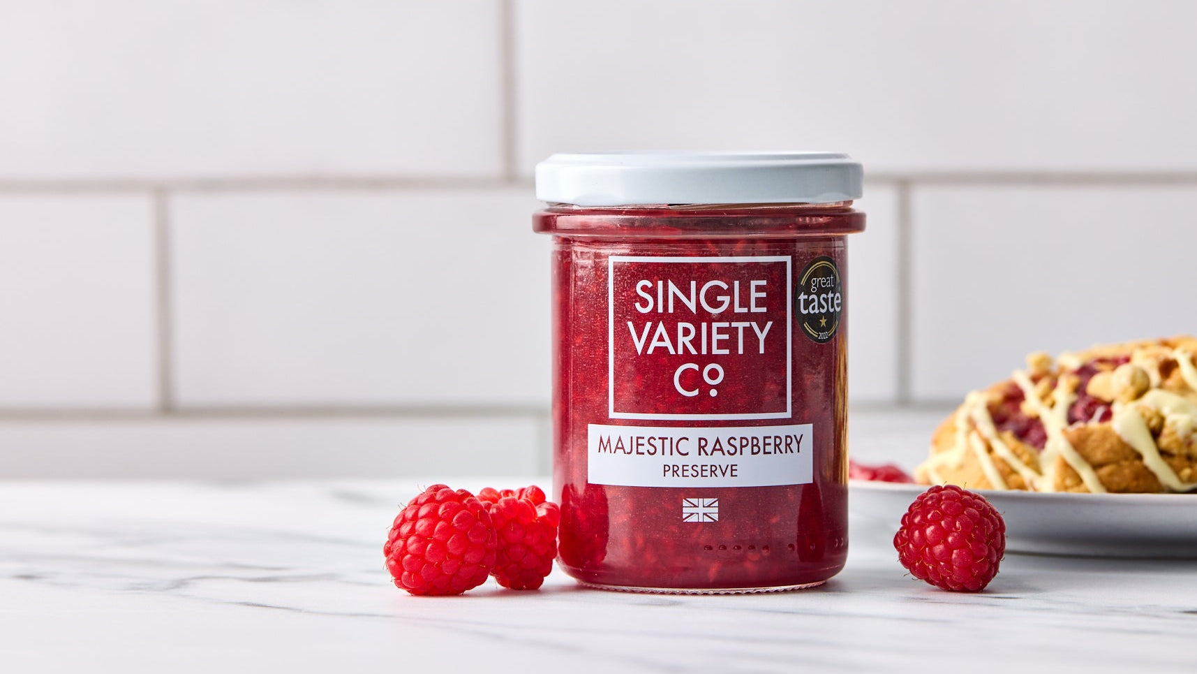 Jar of Single Variety Co. raspberry preserve with raspberries and a pastry on a white surface.